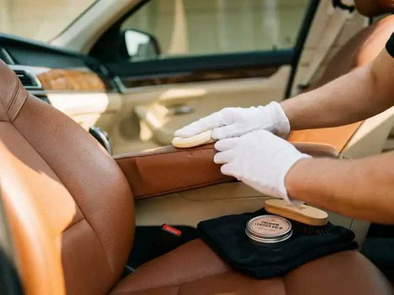Handen in witte handschoenen brengen leeronderhoud aan op cognackleurige autostoelen met gouden lichtinval