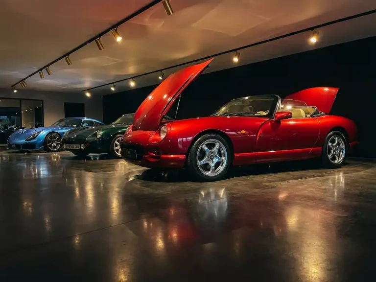 Drie TVR sportwagens in diagonale opstelling in showroom, rode TVR Chimaera op voorgrond met dramatische verlichting